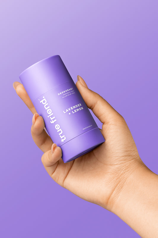 A hand holding a stick of True Friend lavender + lemon deodorant against a purple background