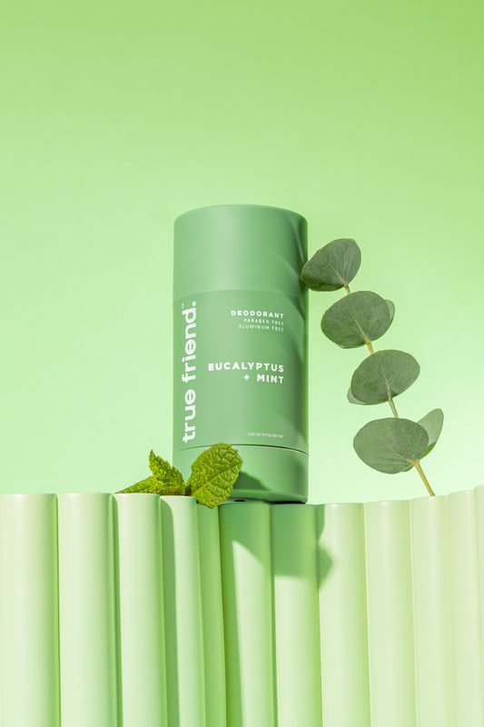 A green deodorant stick labeled 'Eucalyptus Mint' from the brand 'True Friend', displayed upright with a eucalyptus leaf and mint leaves in the foreground.