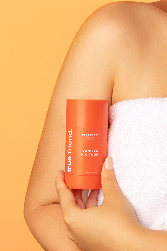 A person's arm holding a stick of deodorant with the brand 'true friend' and the scent 'Vanilla + Citrus' printed on it.