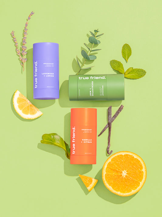 An assortment of personal care products from True Friend, including a deodorant stick, a bar of soap, and a natural Konjac sponge, displayed on a green background with orange slices and greenery.