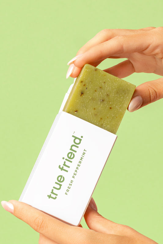 A hand holding a peppermint soap bar that is wrapped in white packaging with the brand name 'True Friend' printed on it.