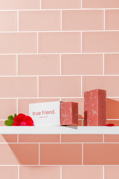 Two pink clay True Friend soap bars sitting on a bathroom shelf with pink tile background