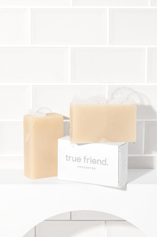 Three organic unscented soap bars in a neutral color, placed on a surface with a white tiled background and packaging box labeled 'TRUE FRIEND'.