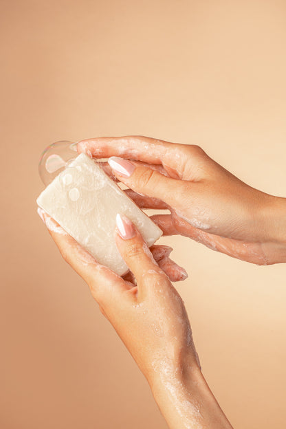 two hands holding a white True Friend Unscented soap bar with some bubbles on a tan background