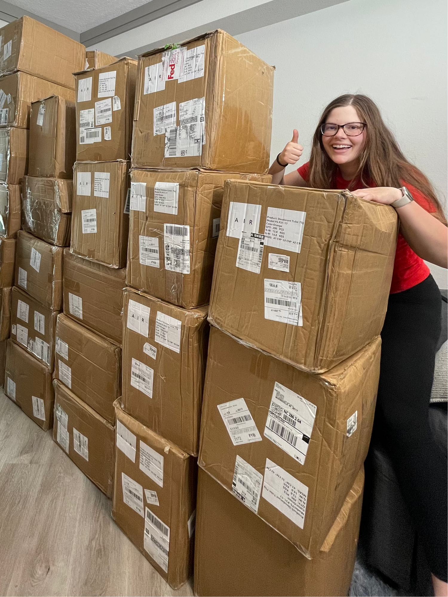 a woman standing next to a large stack of cardboard boxes with shipping labels in a room.